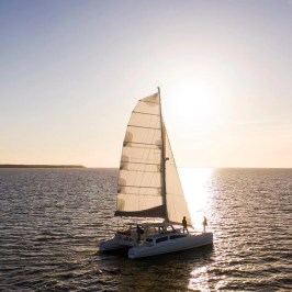 Sailboat catamaran on calm water at sunset, with sun low in the sky.