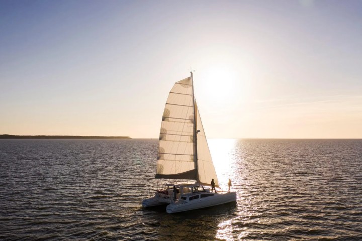 Sailboat catamaran on calm water at sunset, with sun low in the sky.