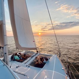People relaxing on a catamaran sailboat at sunset with sails and ocean view.
