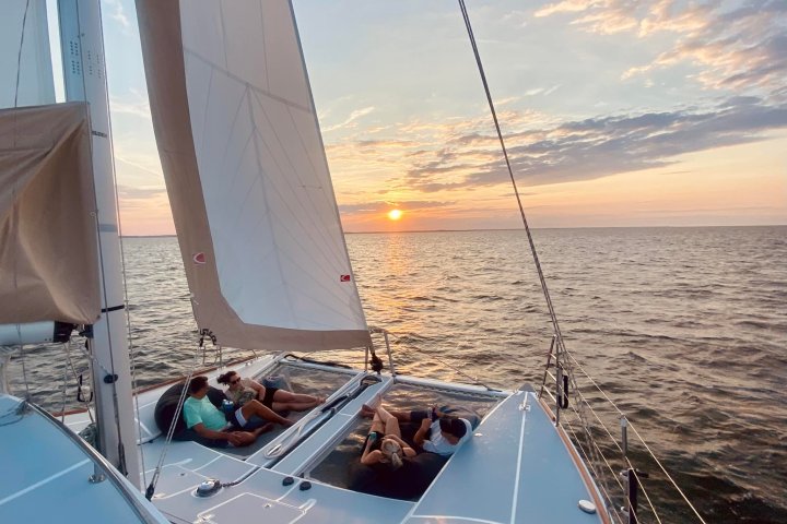 People relaxing on a catamaran sailboat at sunset with sails and ocean view.