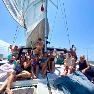 bachelorette party on a catamaran deck, smiling and holding drinks under a clear sky.