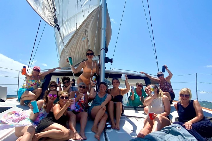 bachelorette party on a catamaran deck, smiling and holding drinks under a clear sky.