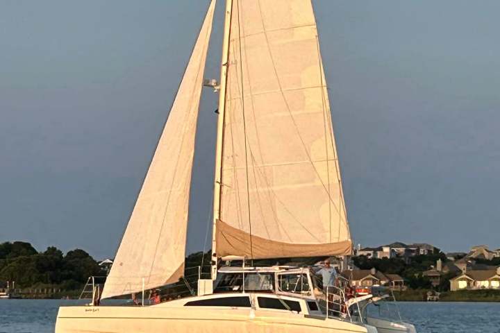 Sailing catamaran on calm water near a residential shoreline at sunset.