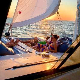 People relaxing on a catamaran sailboat during sunset with sails up and ocean in the background.