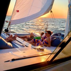 People relaxing on a catamaran sailboat during sunset with sails up and ocean in the background.