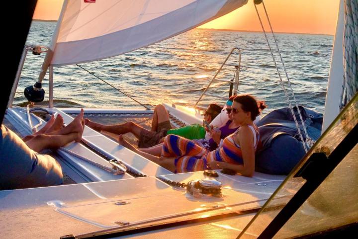 People relaxing on a catamaran sailboat during sunset with sails up and ocean in the background.