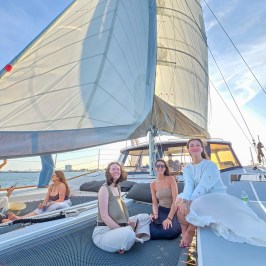 Three people sitting on a sailboat netting with large sails overhead on a sunny day.