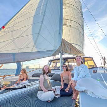 Three people sitting on a sailboat netting with large sails overhead on a sunny day.