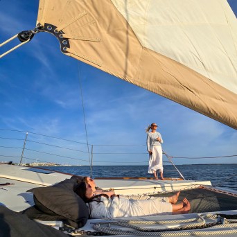 Two people relaxing on a sailboat; one lying down, another standing, with a blue sky and water in the background.