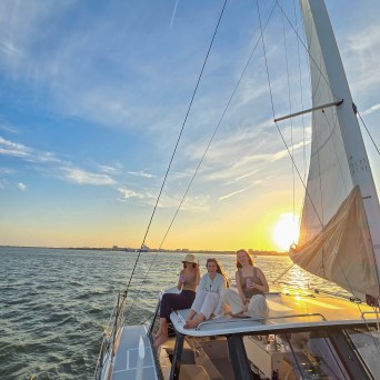 Three people relaxing on a sailboat at sunset over calm water.