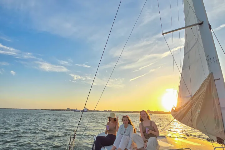 Three people relaxing on a sailboat at sunset over calm water.