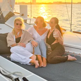 Three people relaxing on a sailboat at sunset, sitting on a netted deck, with a city skyline in the background.