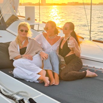Three people relaxing on a sailboat at sunset, sitting on a netted deck, with a city skyline in the background.