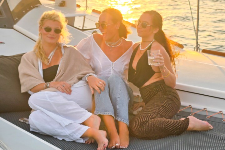 Three people relaxing on a sailboat at sunset, sitting on a netted deck, with a city skyline in the background.