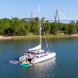 Catamaran anchored in a river with people on deck and colorful inflatables in the water.
