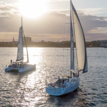 Two sailboats on water at sunset with cityscape in background.