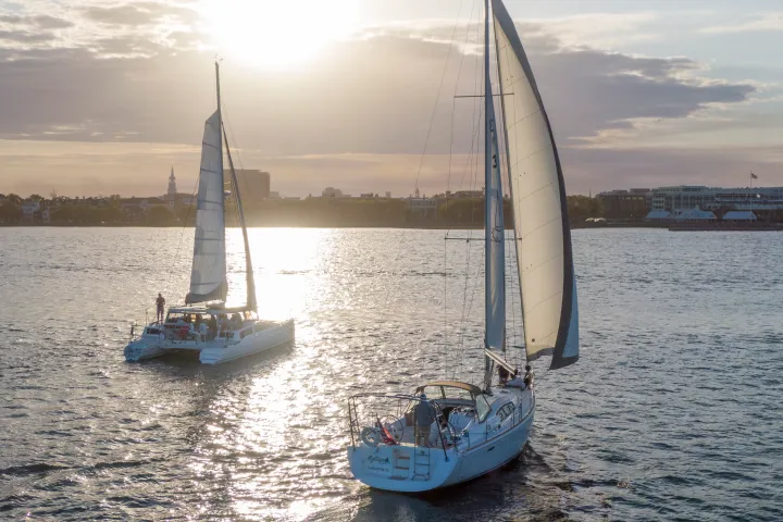 Two sailboats on water at sunset with cityscape in background.