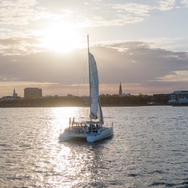 Sailboat on water with setting sun and city skyline in background.