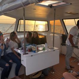 Group of people sitting on a boat at sunset, with snacks on a table.