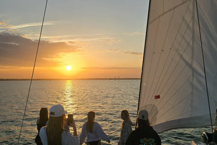 People on a sailboat watch the sunset over the ocean.