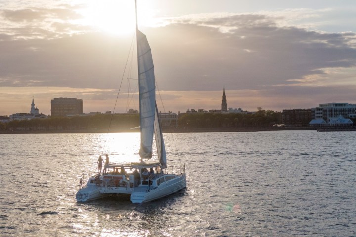 Catamaran sailing at sunset with city skyline in background.