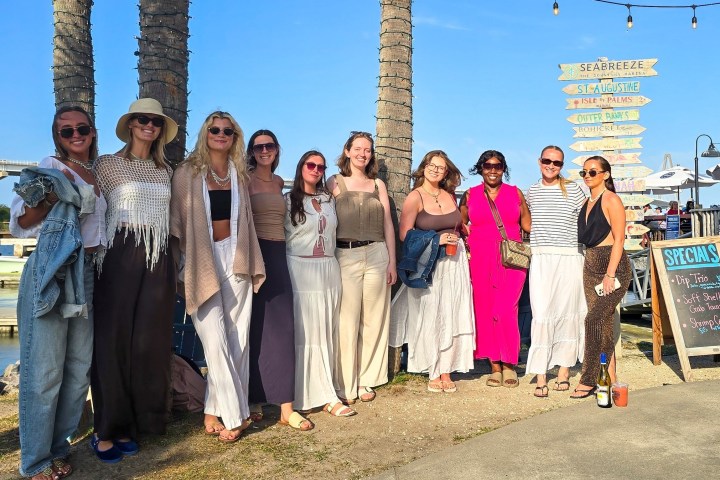 Group of women posing outdoors near palm trees and signpost by water.