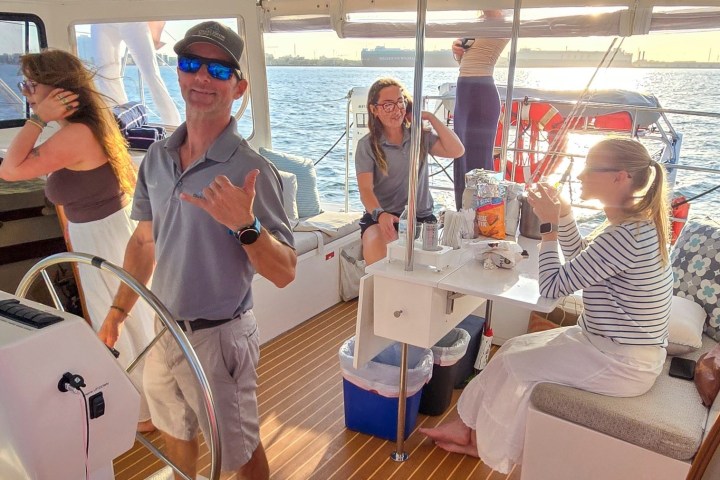 People relaxing and talking on a boat, one steering, with a sunny ocean backdrop.